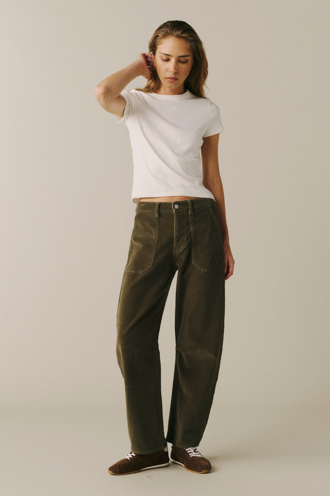 A woman modeling a pair of dark olive green wide-leg corduroy pants, styled with a white t-shirt and brown shoes, against a plain beige studio background.