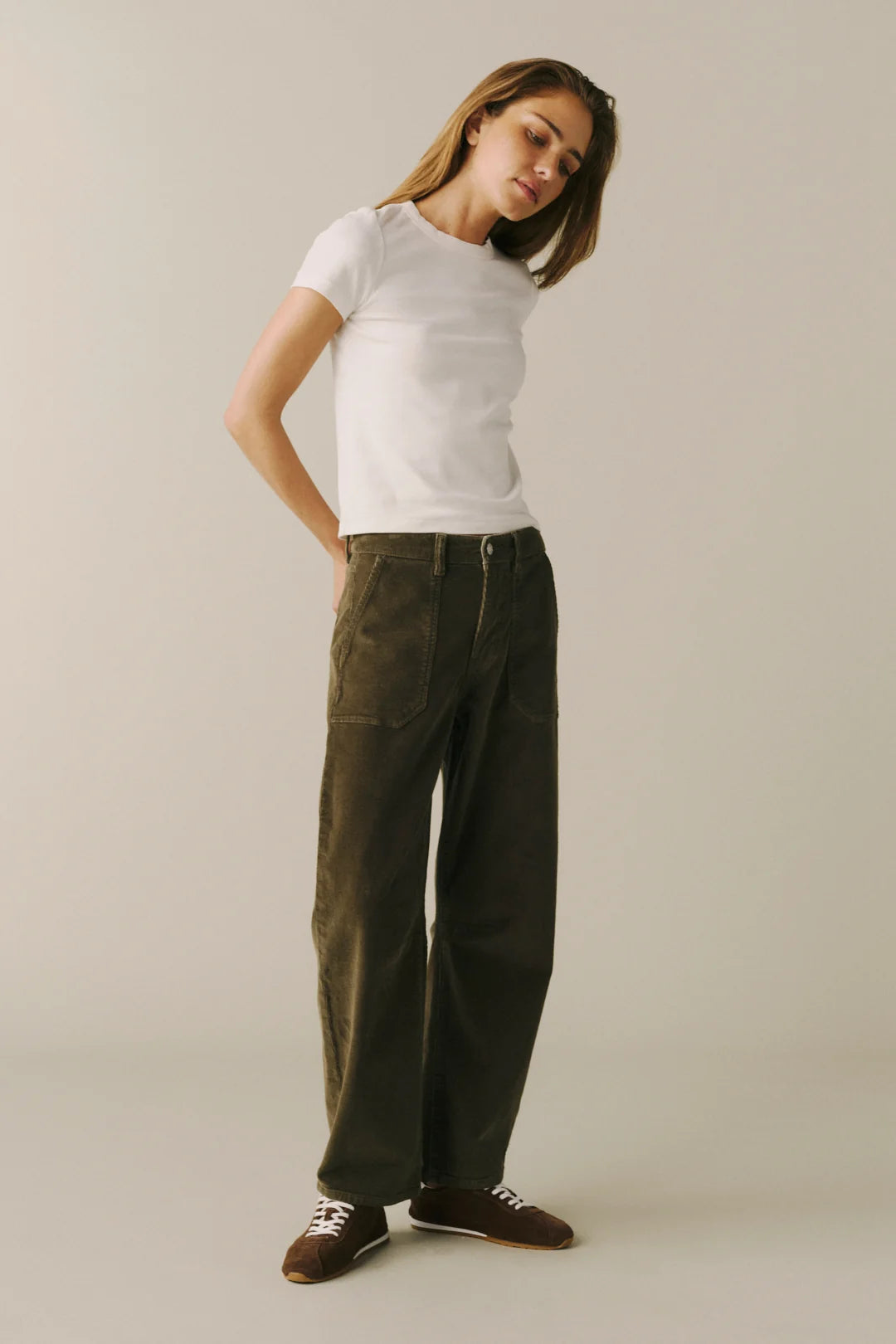 A woman modeling dark olive green wide-leg corduroy pants, styled with a white t-shirt and brown sneakers, against a plain light beige studio background.