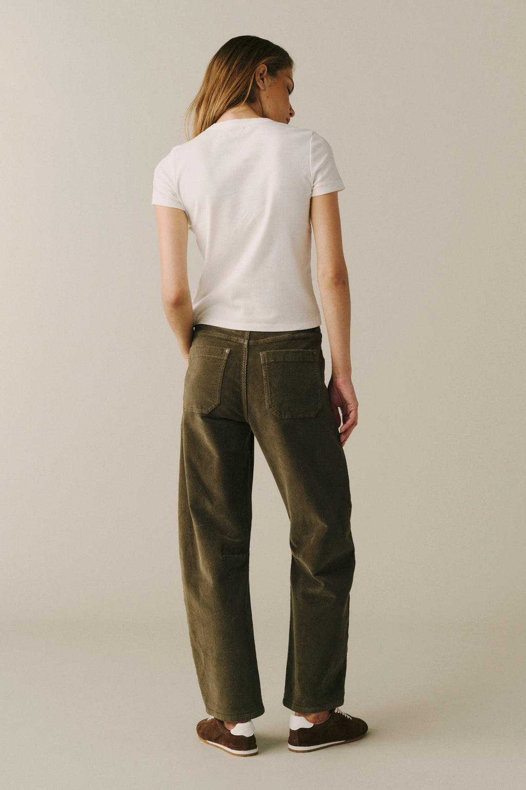 A woman shown from the back wearing a white short-sleeve t-shirt, olive green wide-leg corduroy pants, and brown shoes, standing against a plain beige studio background.