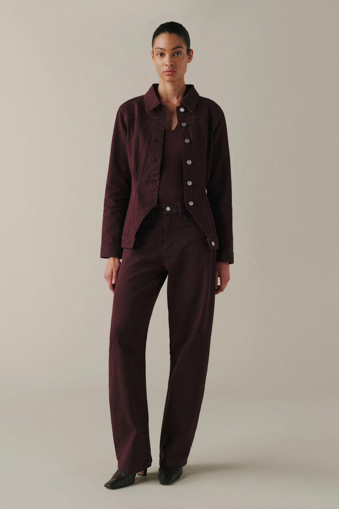 A model stands against a plain light gray studio background wearing a matching dark burgundy button-front jacket and trousers set with black pointed shoes.