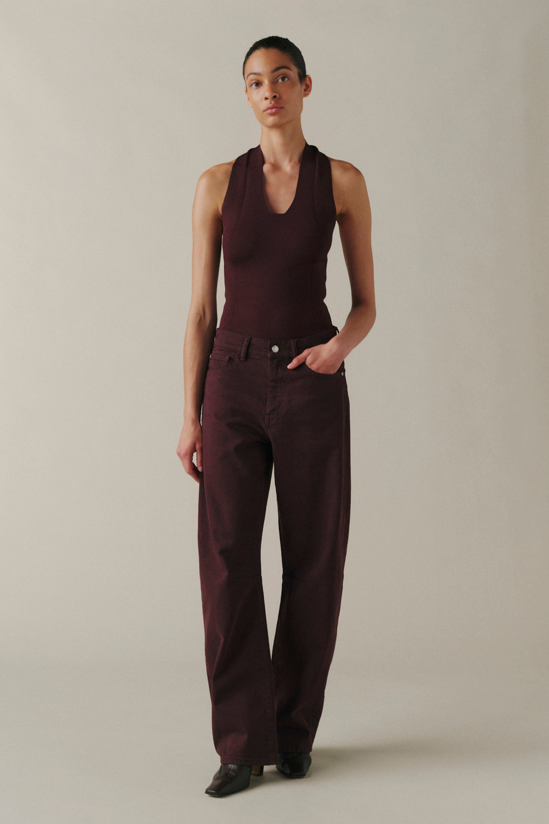 A model stands against a plain beige studio background wearing a sleeveless dark burgundy top and matching wide-leg trousers, with black shoes.