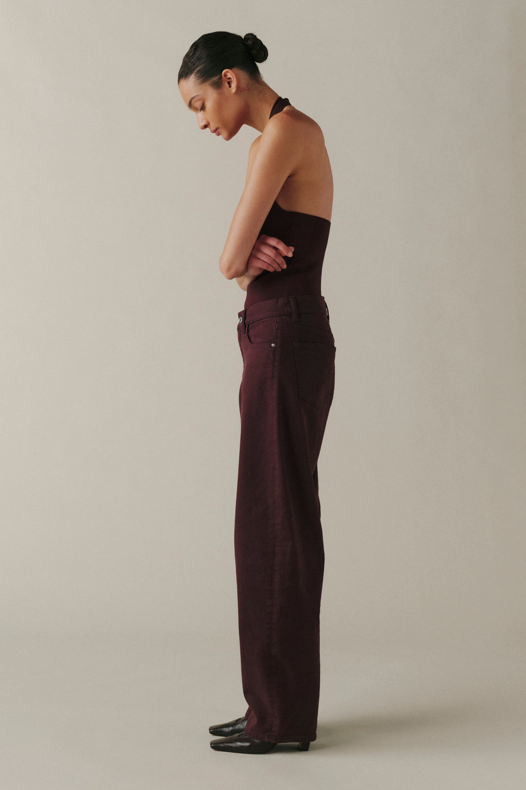 A woman in a side profile wearing a dark burgundy strapless top and matching wide-leg trousers, standing against a plain beige studio background.