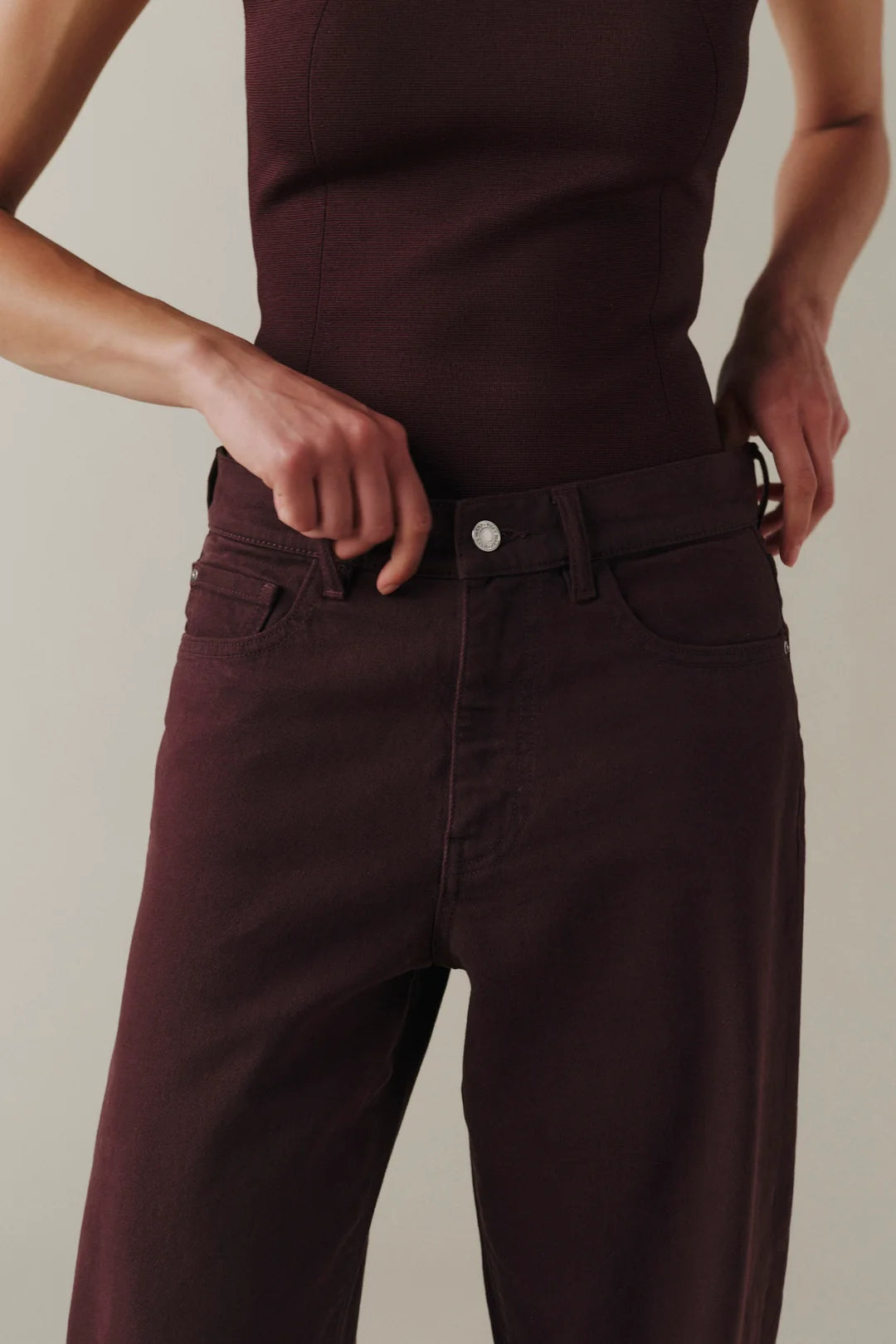 Close-up of a person wearing dark plum-colored denim jeans and a matching dark top, with hands adjusting the waistband.
