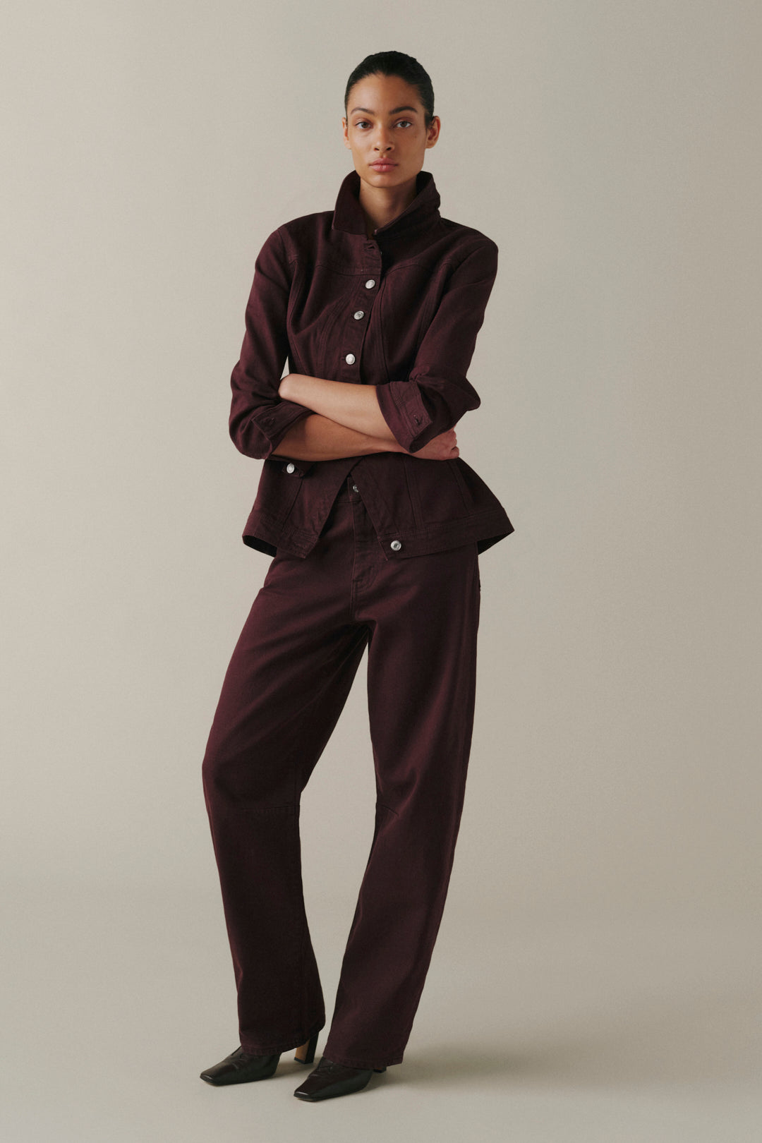 A model stands against a plain beige studio background wearing a matching dark burgundy button-front jacket and trousers set.