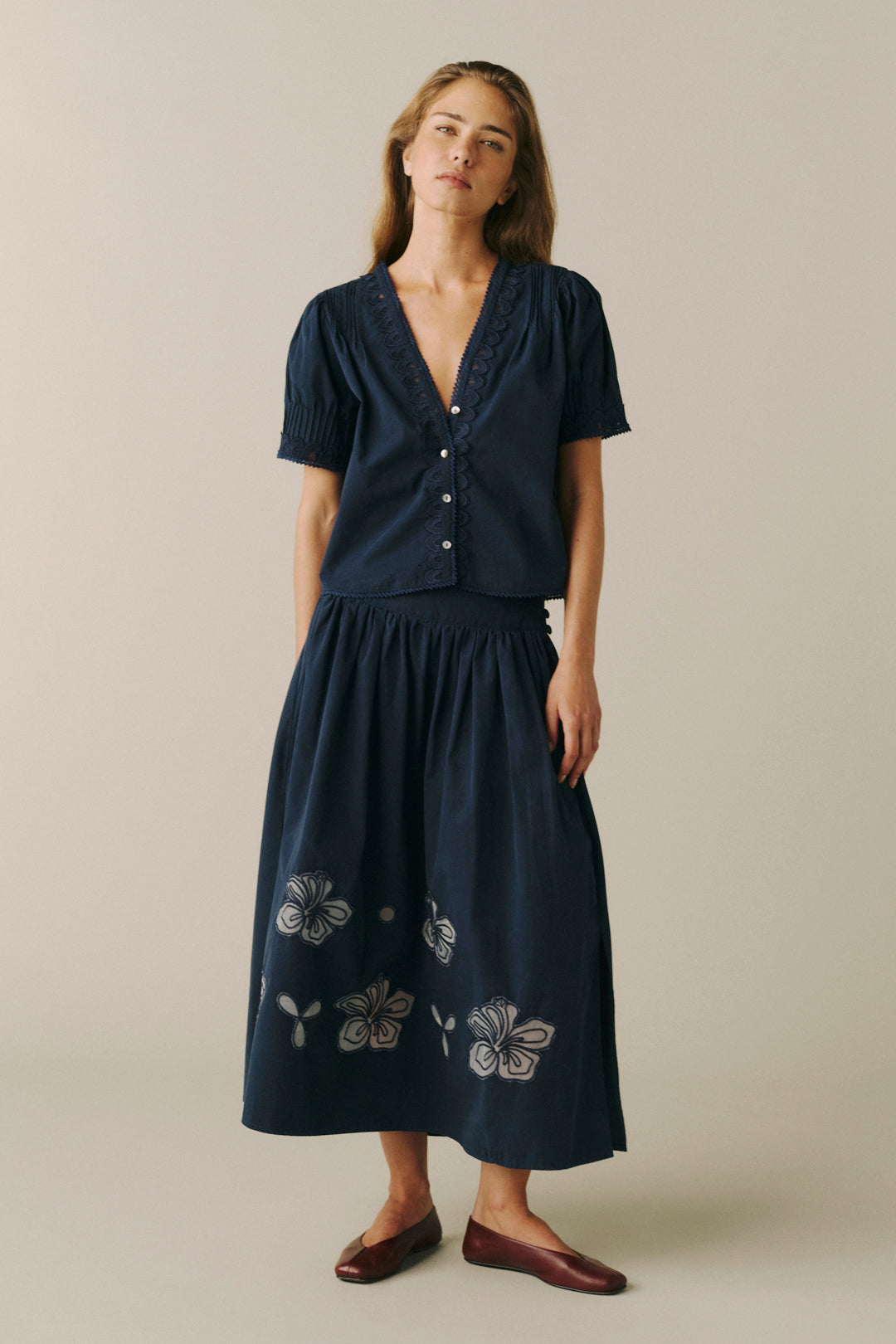 A woman modeling a dark navy blue two-piece set consisting of a short-sleeve button-front top and a matching midi skirt with white floral embroidery near the hem, paired with dark brown flats, against a solid light beige background.