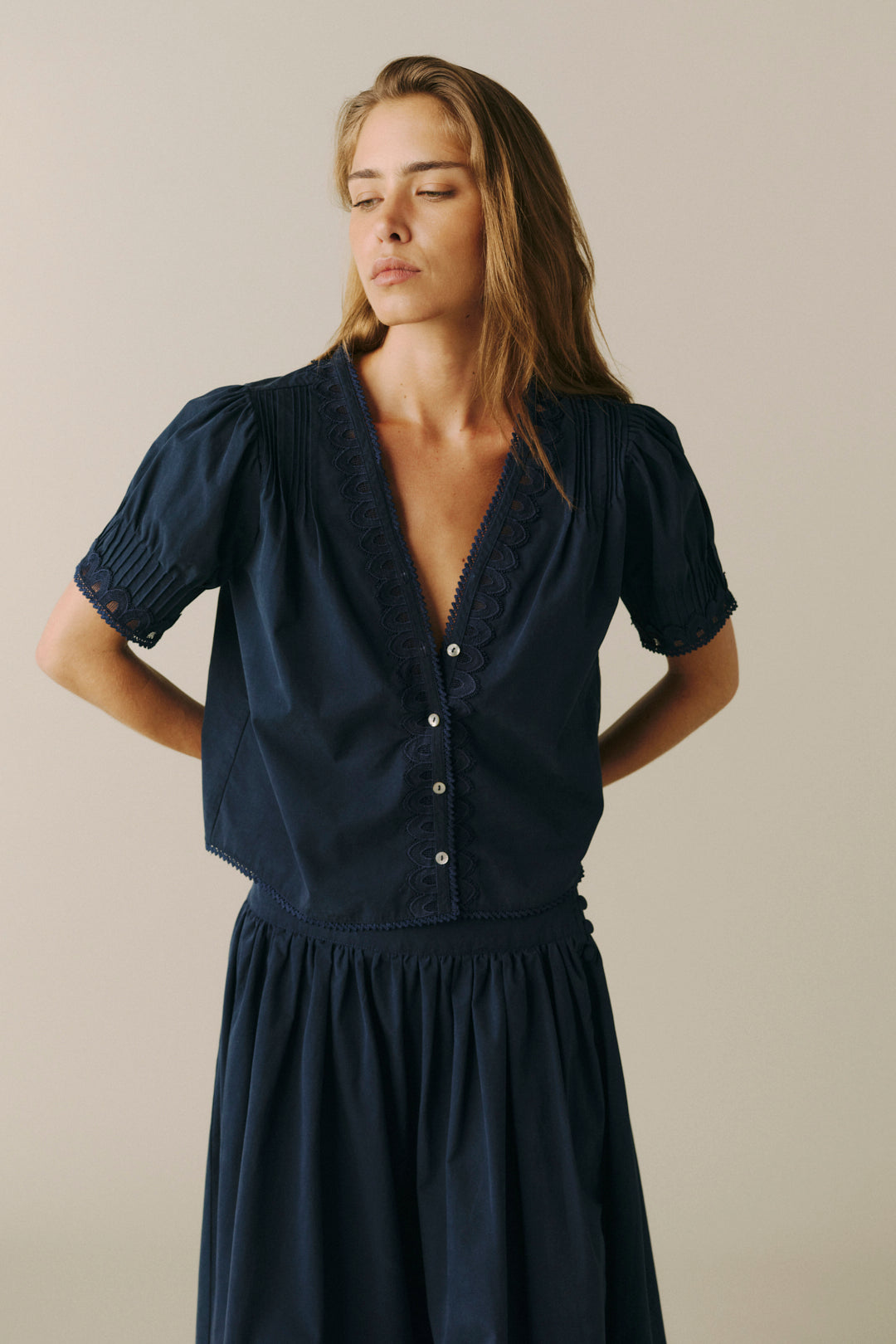 A woman wearing a dark navy blue short-sleeve blouse with puff sleeves, a buttoned front, and lace trim along the V-neck, paired with a matching navy skirt, set against a plain beige studio background.