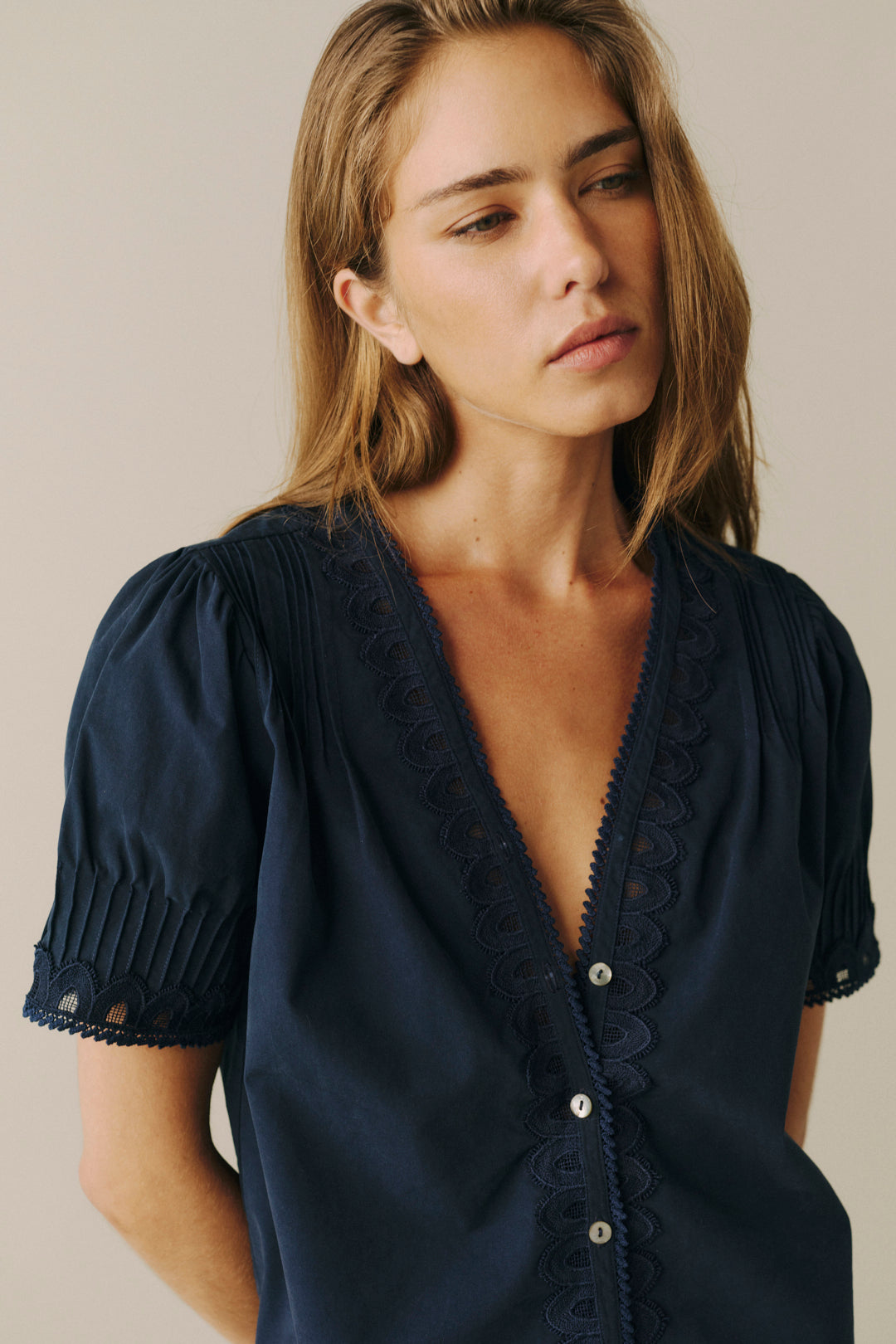 A woman wearing a navy blue short-sleeve blouse with pleated sleeves, a scalloped lace-trimmed V-neckline, and decorative buttons, photographed against a plain light beige background.