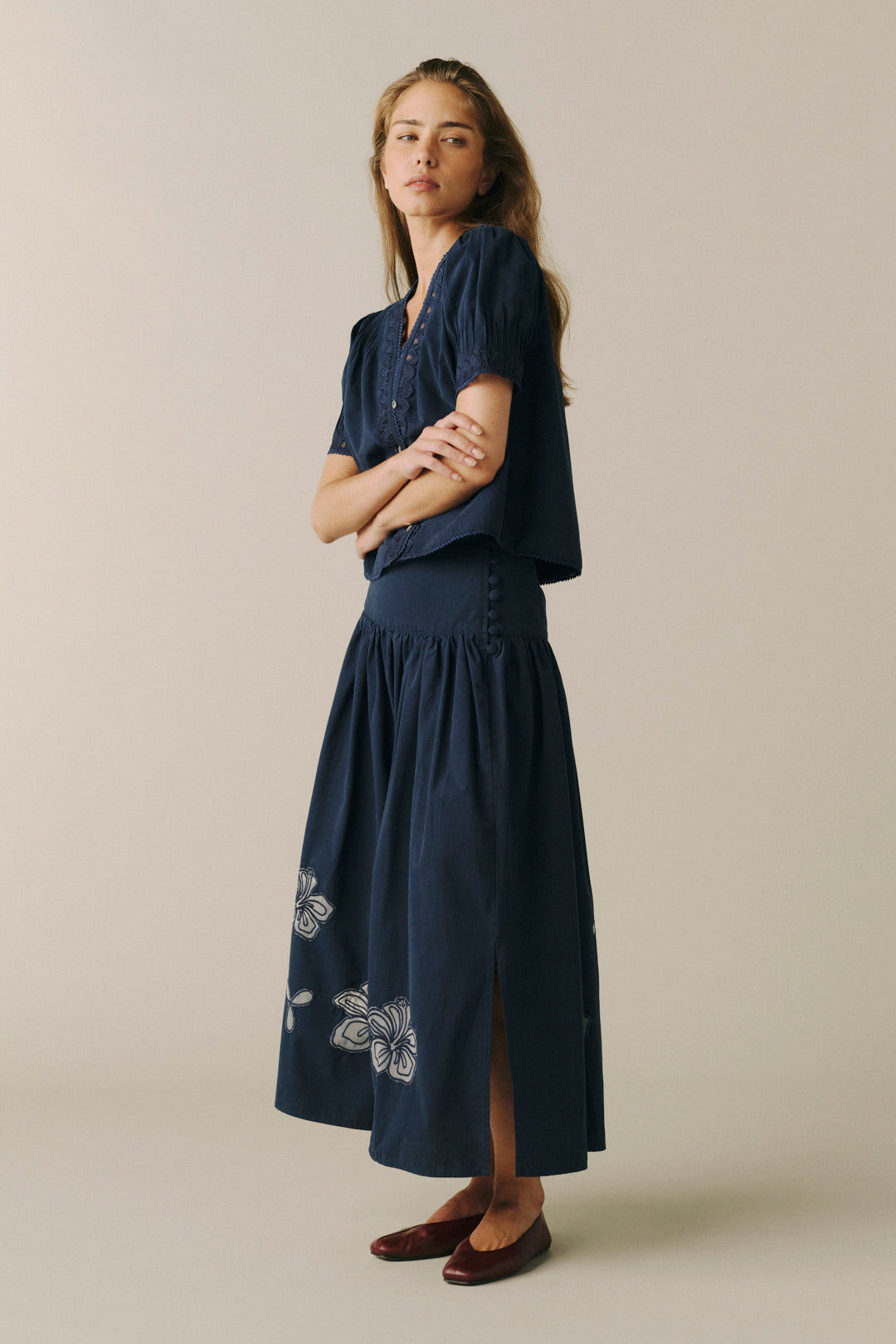 A woman modeling a navy blue two-piece set consisting of a short-sleeve button-front top and a matching high-waisted skirt with white floral embroidery near the hem, paired with dark brown flats, against a solid light beige background.