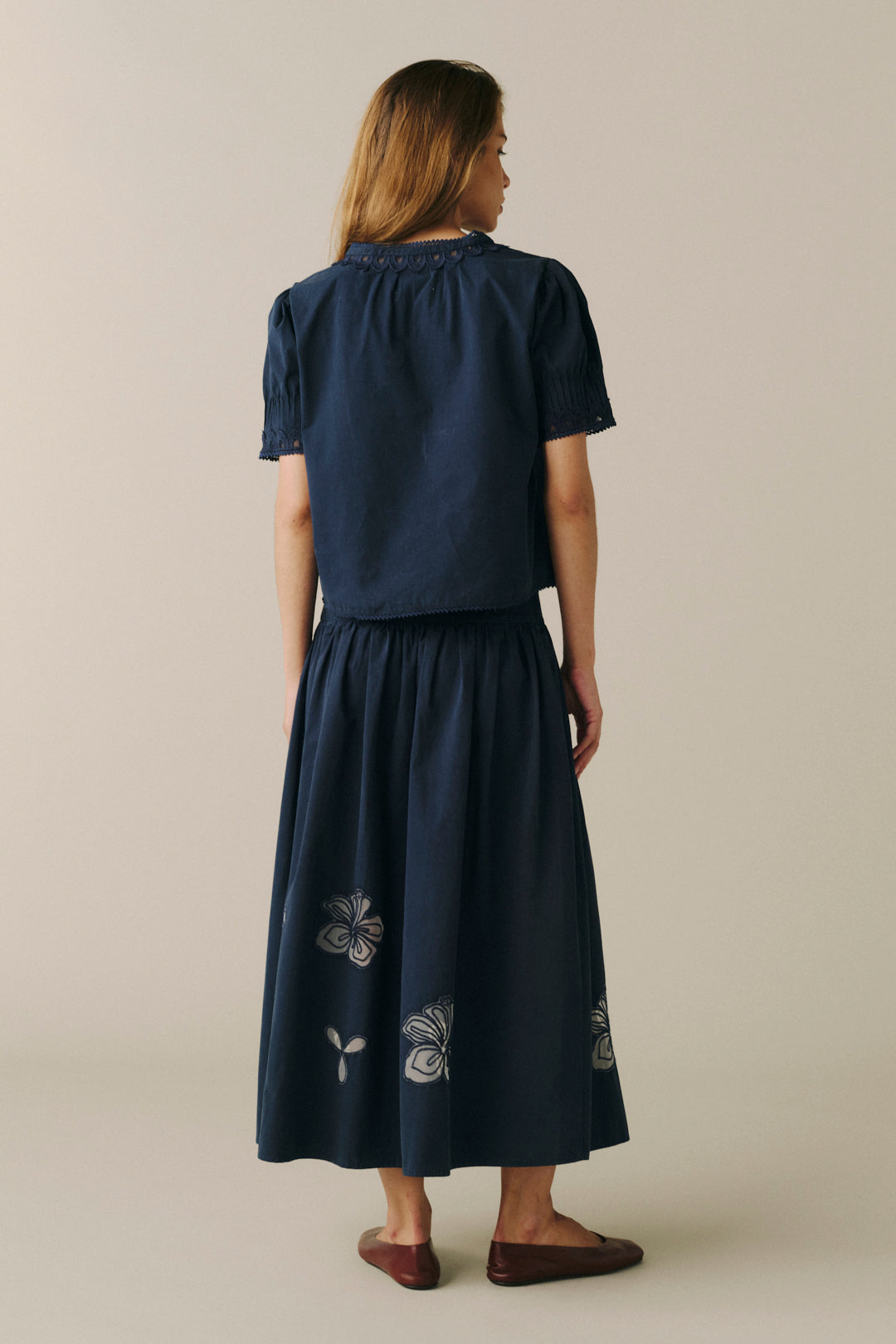 A woman viewed from behind wearing a navy blue two-piece set consisting of a short-sleeve top with a smocked collar and a matching high-waisted A-line skirt with white floral embroidery near the hem, set against a plain beige studio background.