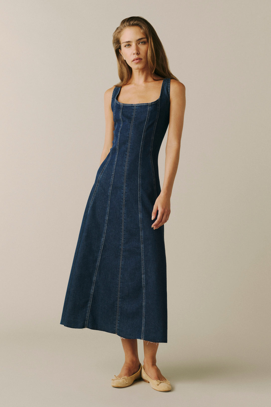 A woman modeling a dark blue denim midi dress with a square neckline and side pockets, paired with beige flat shoes, against a solid light beige studio background.