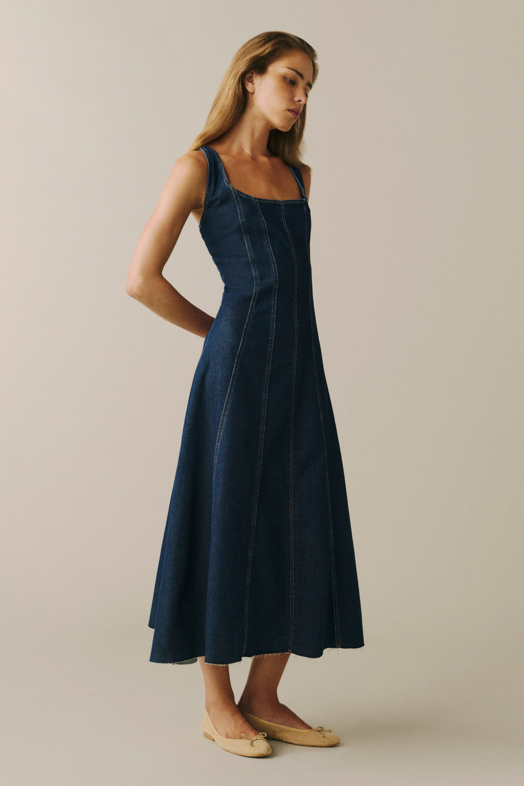 A woman modeling a dark indigo denim midi dress with a square neckline and thin shoulder straps, standing against a solid light beige background.