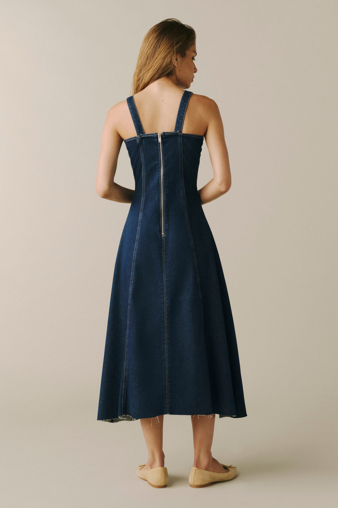 A woman viewed from the back wearing a dark blue denim midi dress with shoulder straps and a visible center back zipper, paired with tan flats, against a plain beige studio background.