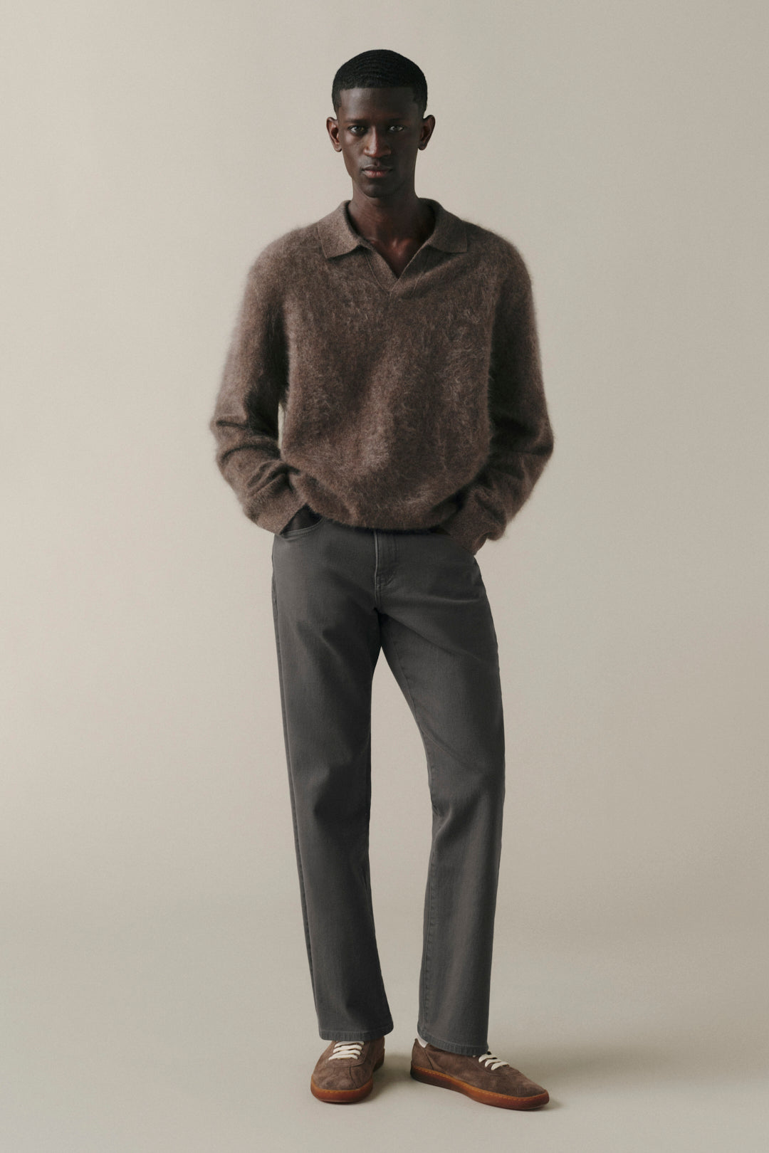A male model stands against a plain warm beige studio background wearing a fuzzy brown knit polo sweater, dark charcoal gray trousers, and brown leather loafers with white stripes.