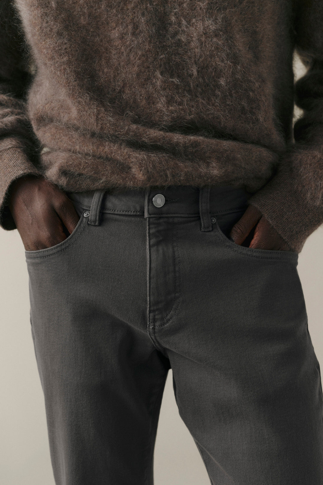 Close-up of a person wearing dark charcoal gray jeans with a brown fuzzy knit sweater, standing with hands in pockets against a light neutral background.