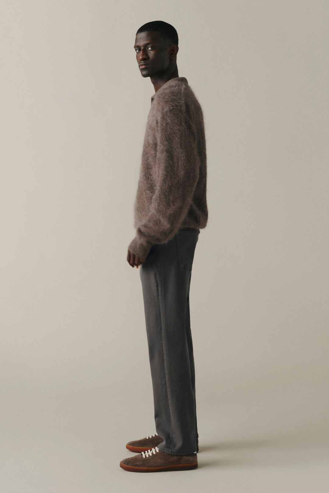 A man stands in profile wearing a fuzzy taupe-brown knit sweater, dark gray trousers, and brown lace-up shoes against a plain warm beige studio background.