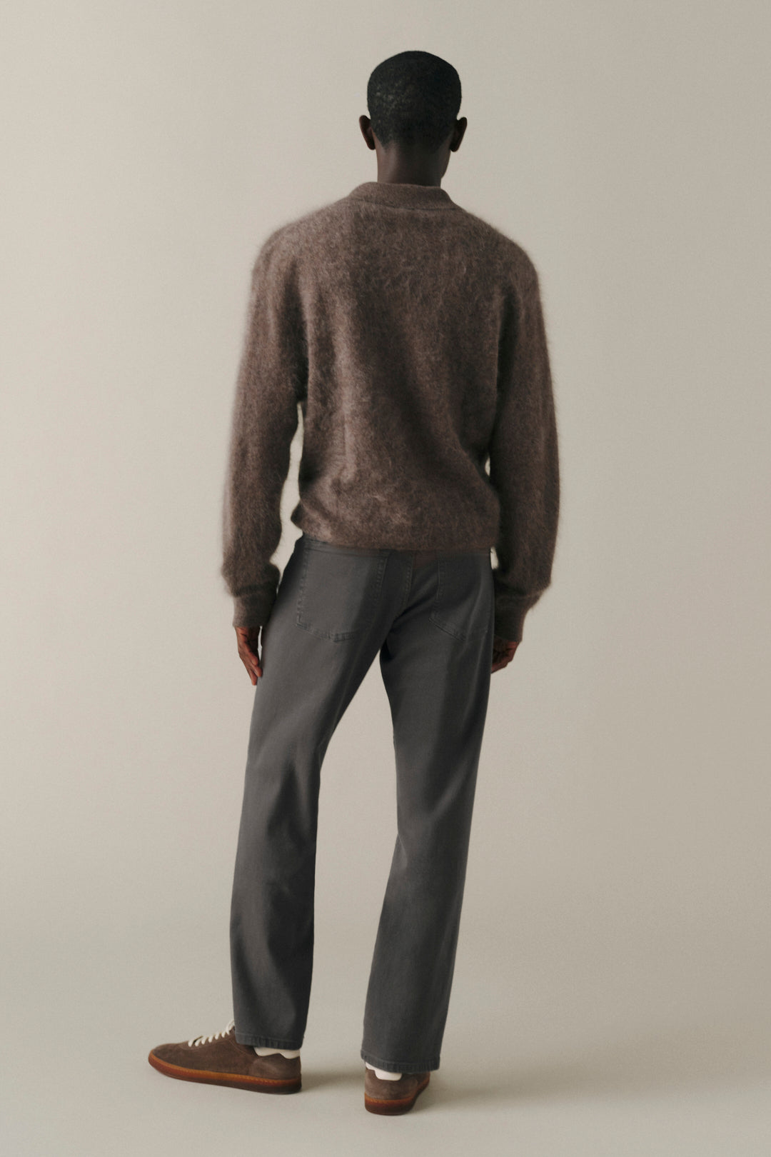 A man shown from the back wearing a brown fuzzy knit sweater, dark gray trousers, and brown shoes, standing against a plain light beige studio background.