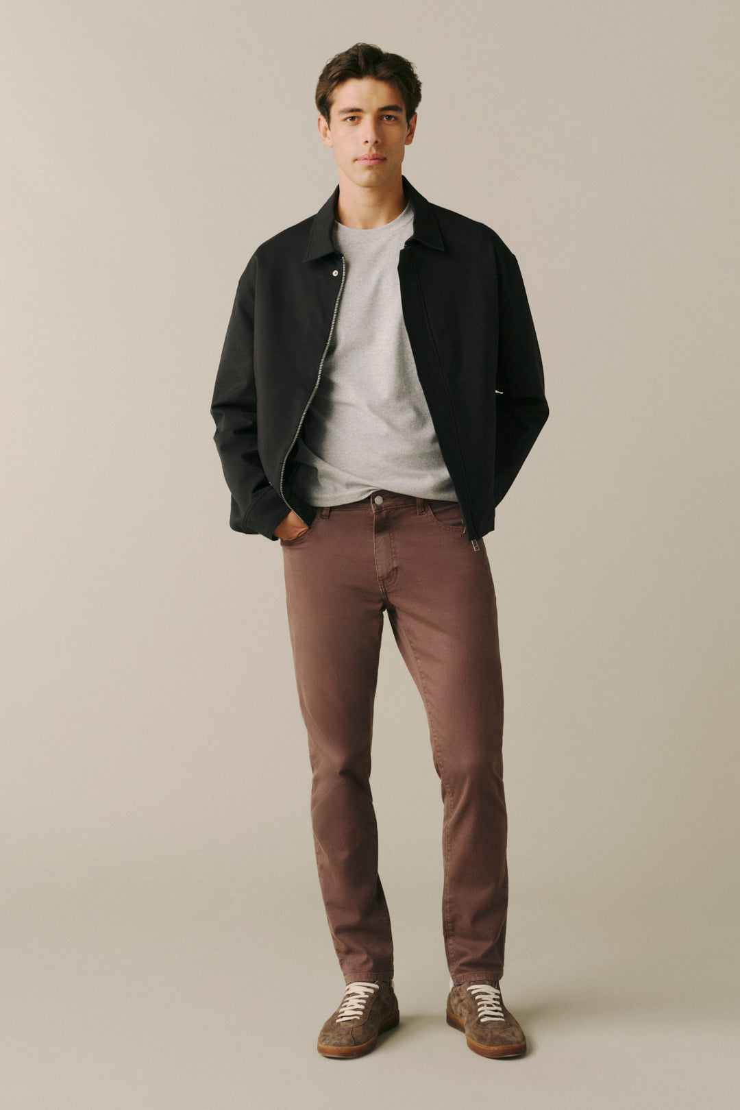 A man modeling a black zip-up jacket over a light gray t-shirt, paired with brown pants and brown suede shoes, standing against a solid light beige background.
