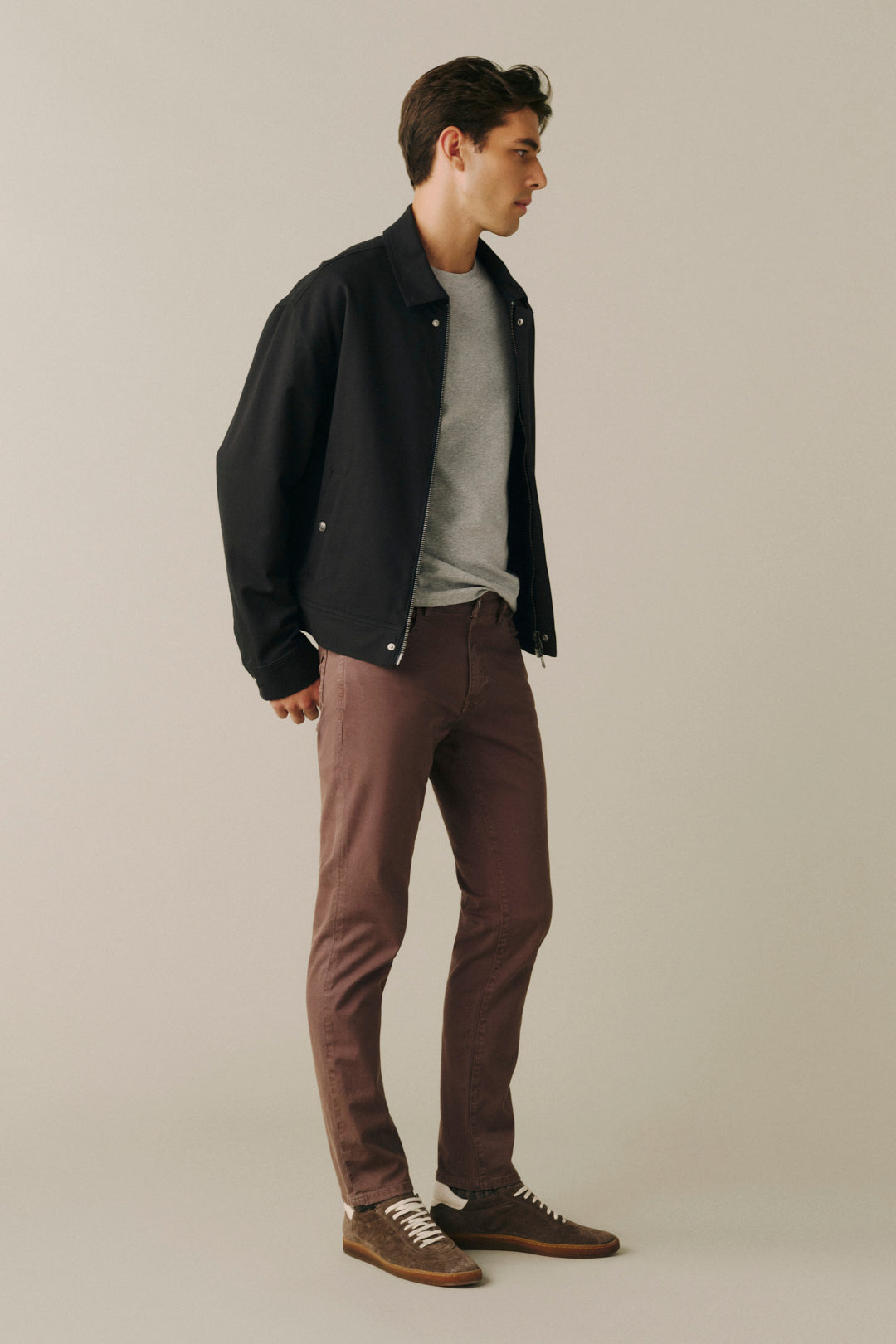 A man wearing a black bomber jacket, gray crewneck t-shirt, brown pants, and brown suede shoes, standing in profile against a solid light beige background.