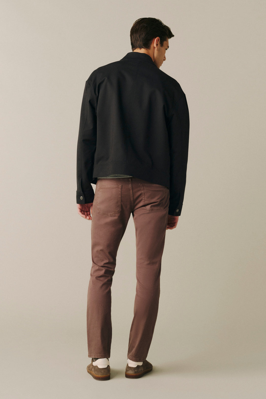 A man shown from behind wearing a black jacket, brown trousers, and brown shoes, standing against a plain light gray studio background.