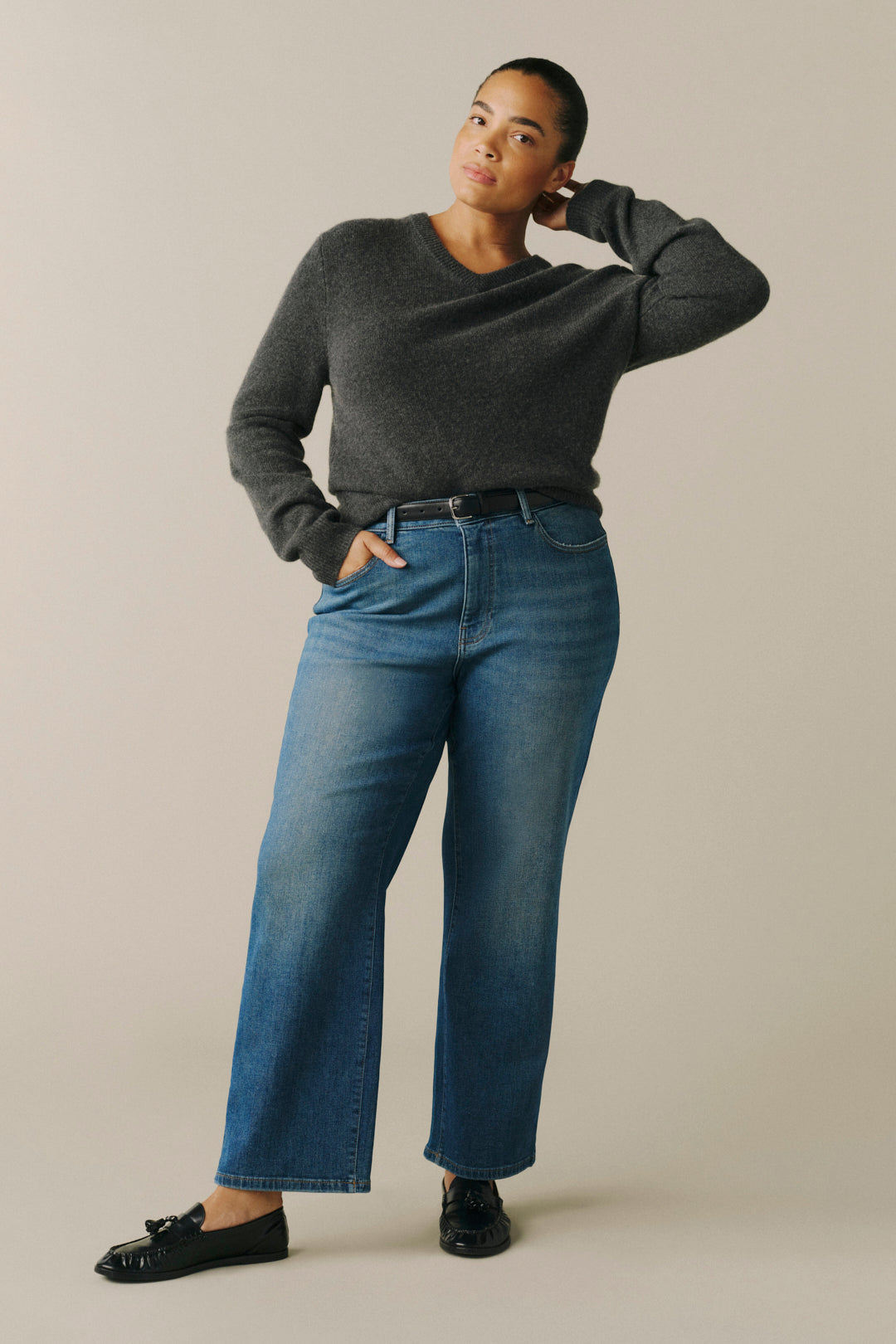 A woman modeling a dark gray long-sleeve knit sweater and blue wide-leg jeans with black loafers.
