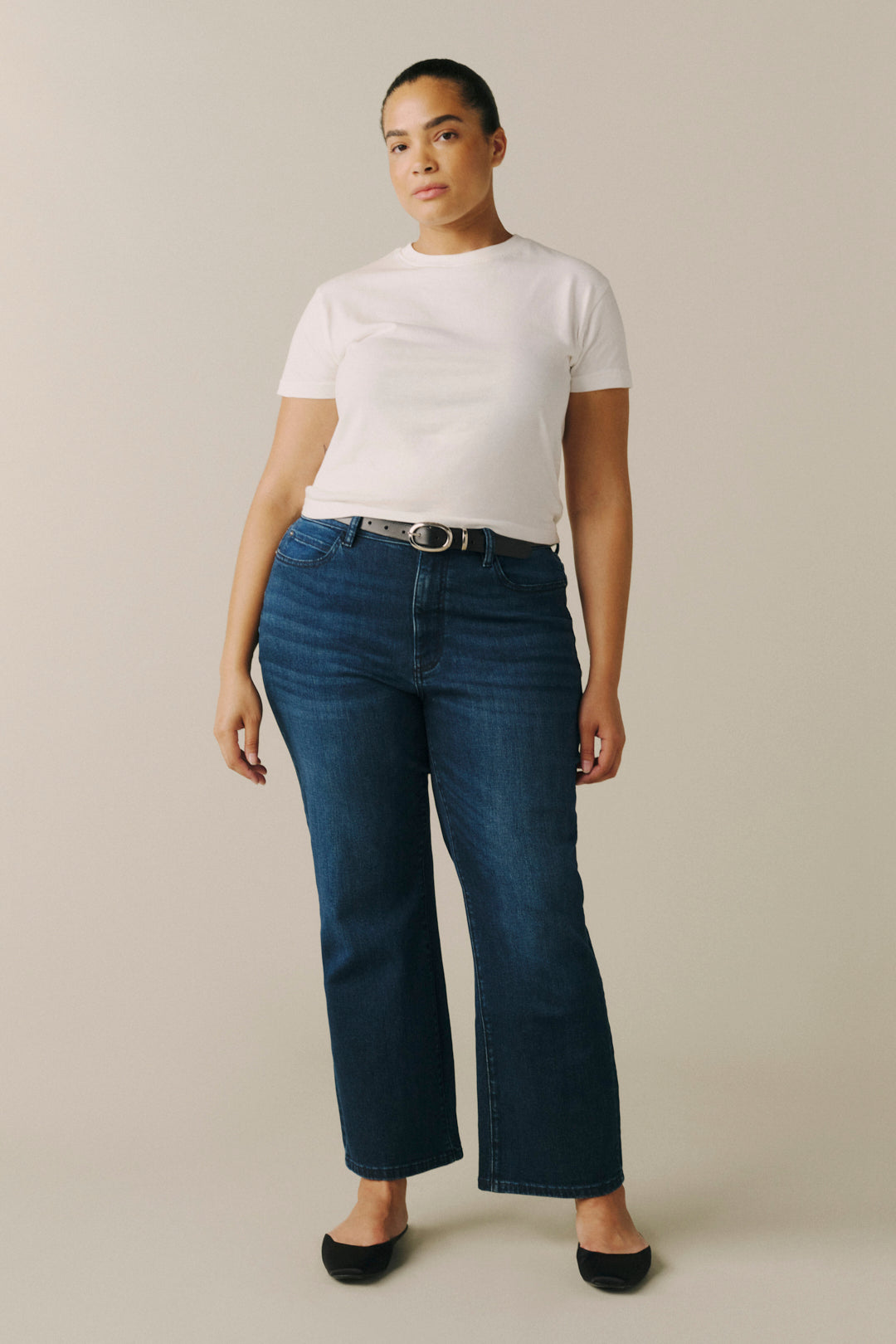 A model wearing dark blue cropped flare jeans with a silver belt, a white t-shirt, and black flats.