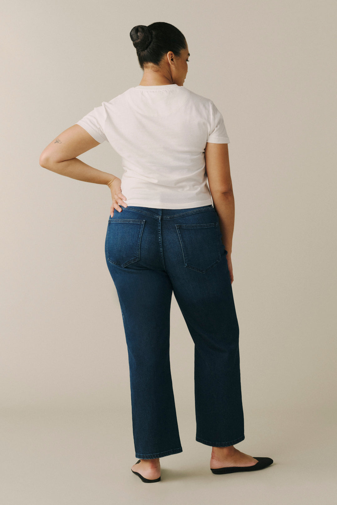 A woman shown from the back wearing dark blue straight-leg jeans, a white t-shirt, and black flats.