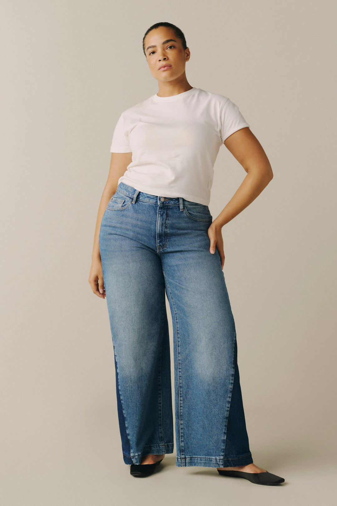 A model wearing a pair of wide-leg blue jeans with a faded wash, styled with a white t-shirt and black flats, standing against a solid beige studio background.