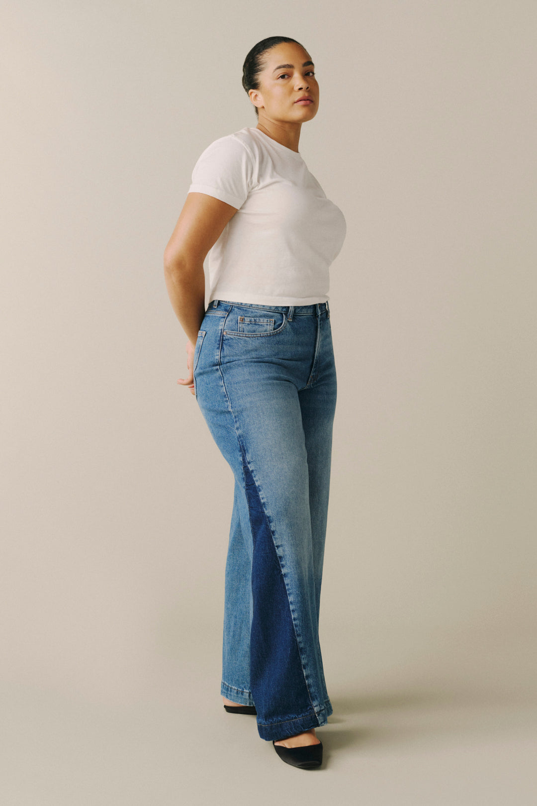 A woman modeling a pair of high-waisted, wide-leg blue jeans with a faded denim wash, paired with a white t-shirt and black flats, standing against a solid light beige background.