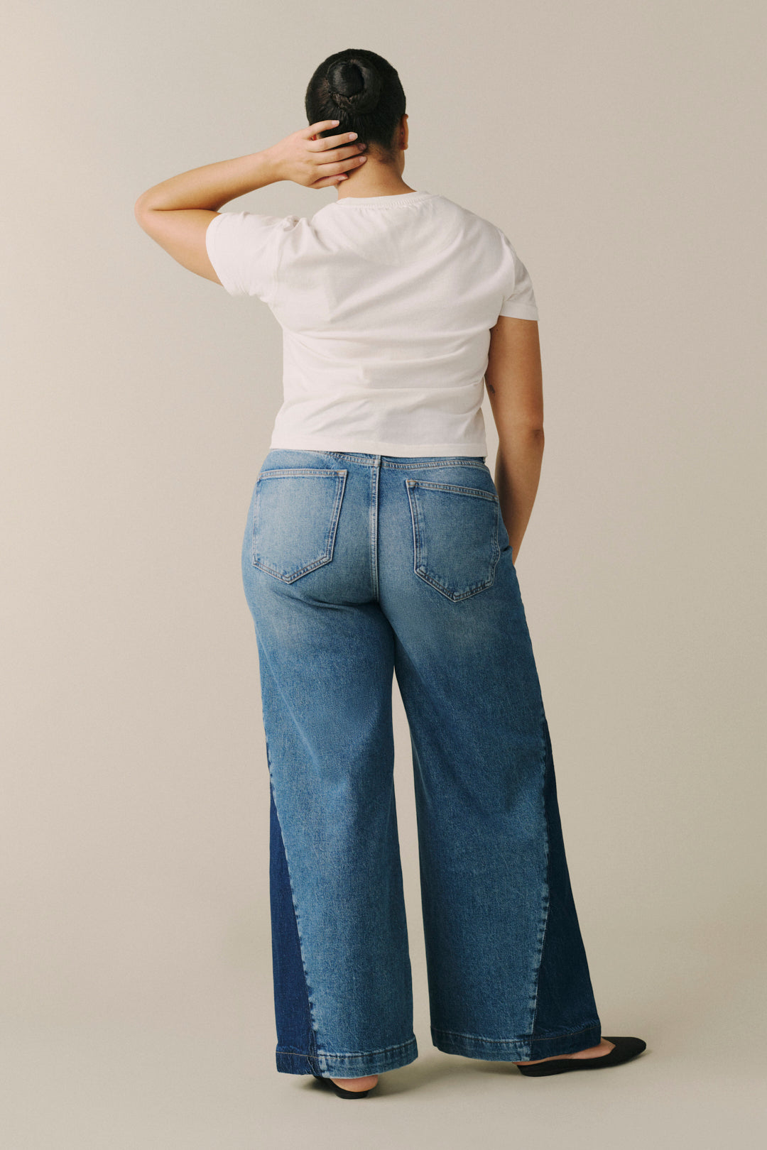 A model shown from the back wearing wide-leg blue denim jeans with dark indigo side panels, a white t-shirt, and black flat shoes, set against a plain beige studio background.
