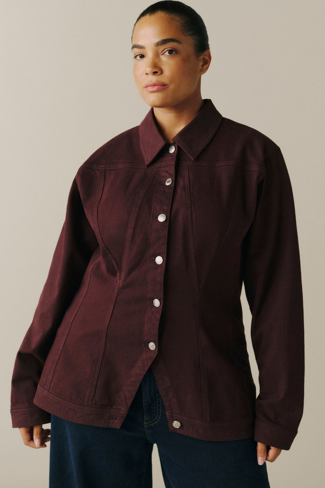 A model wearing a dark burgundy button-up shirt jacket with a pointed collar and silver buttons, paired with dark blue jeans.