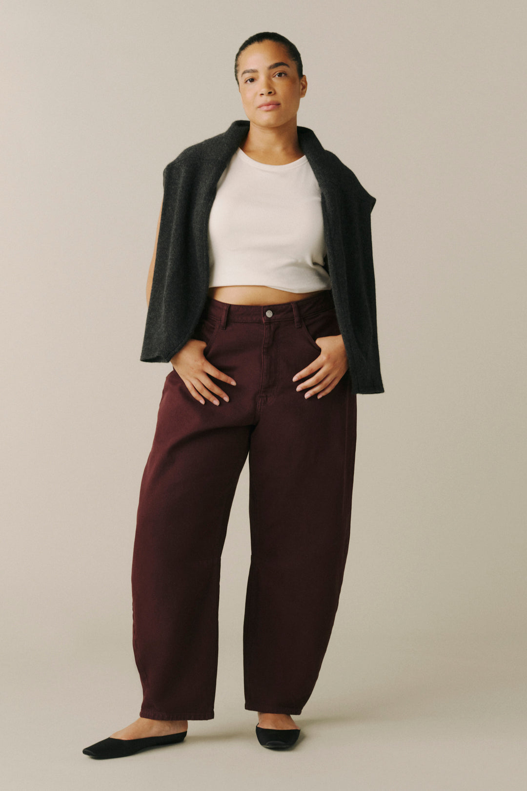 A model wearing dark burgundy wide-leg trousers, a white cropped tank top, a dark charcoal sleeveless cardigan.