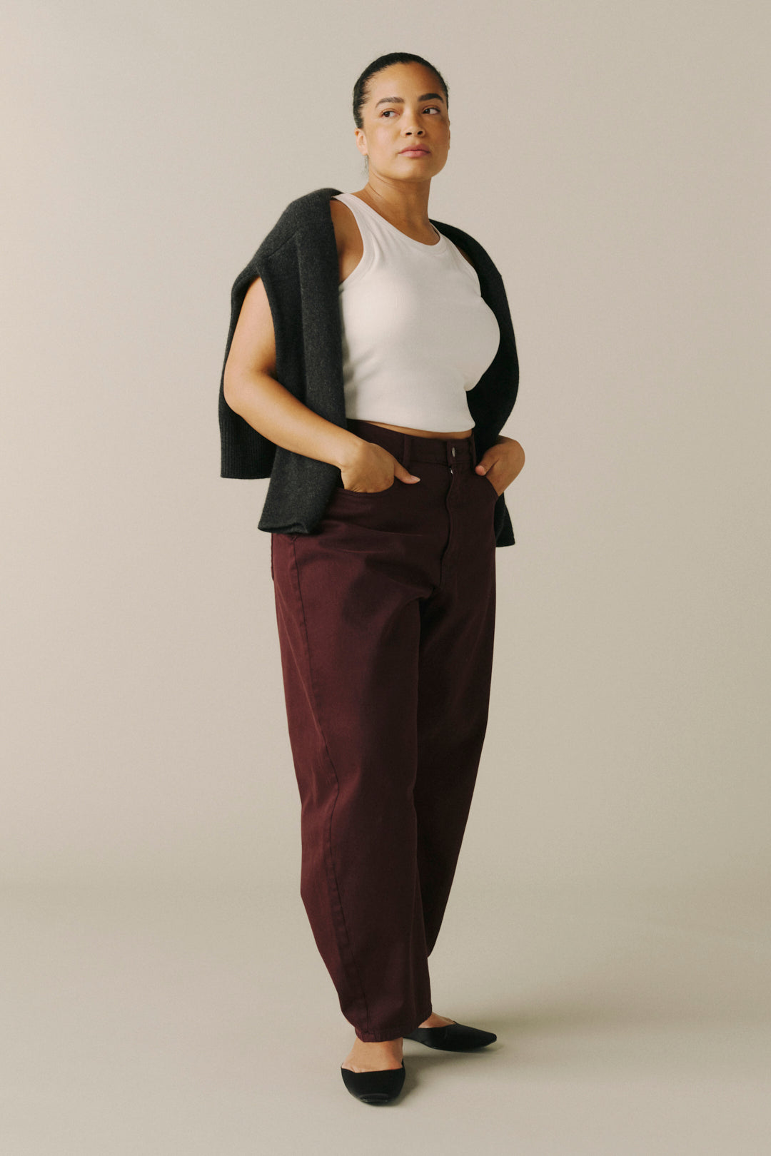 A model stands in a studio wearing a white ribbed tank top, a dark charcoal knit cardigan draped over her shoulders.