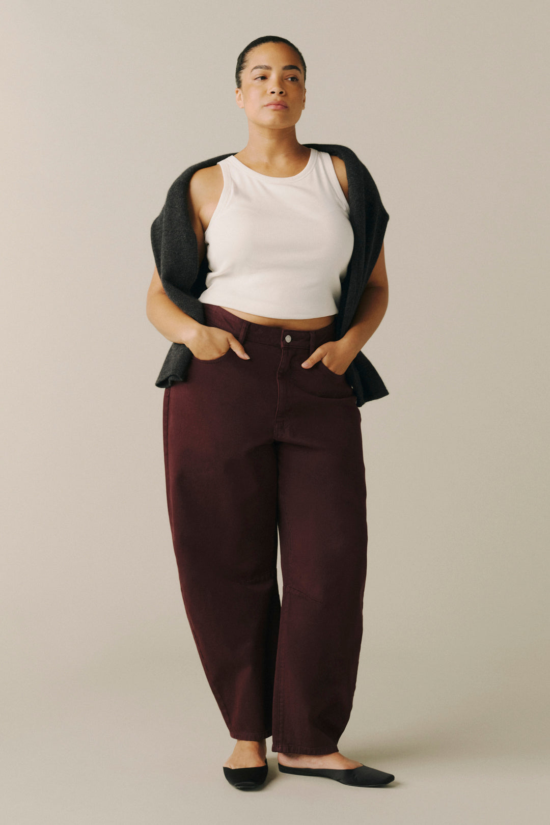 A model wearing dark burgundy wide-leg pants, a white ribbed tank top, a black cardigan draped over her shoulders.
