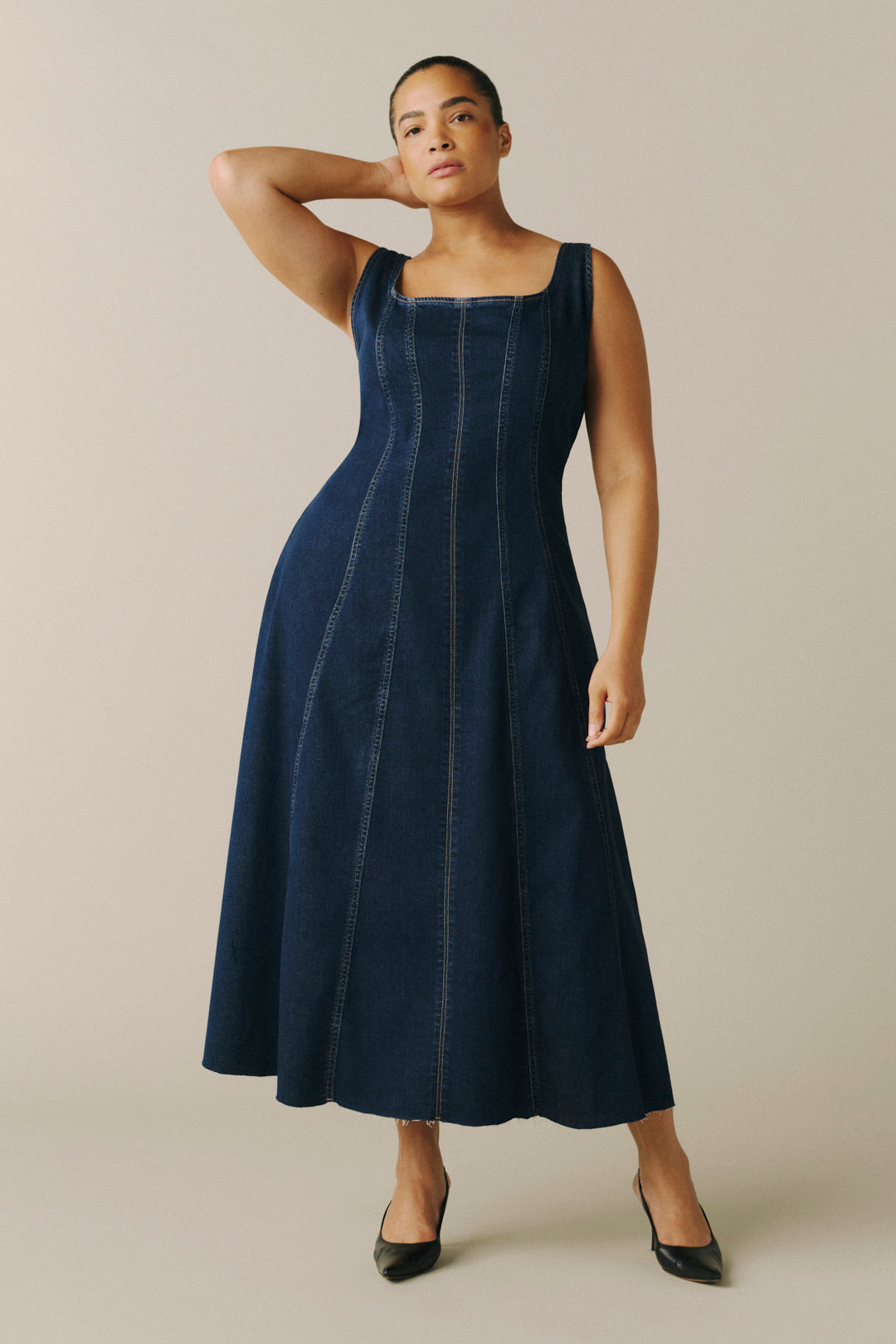 A woman modeling a sleeveless dark blue denim midi dress with vertical seam detailing.
