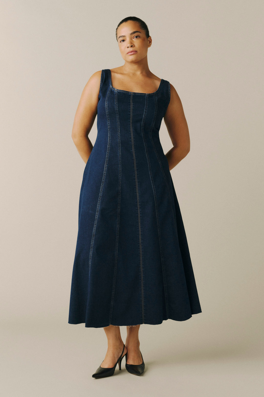 A woman modeling a dark indigo denim midi dress with a square neckline, thick shoulder straps, and vertical seam detailing.