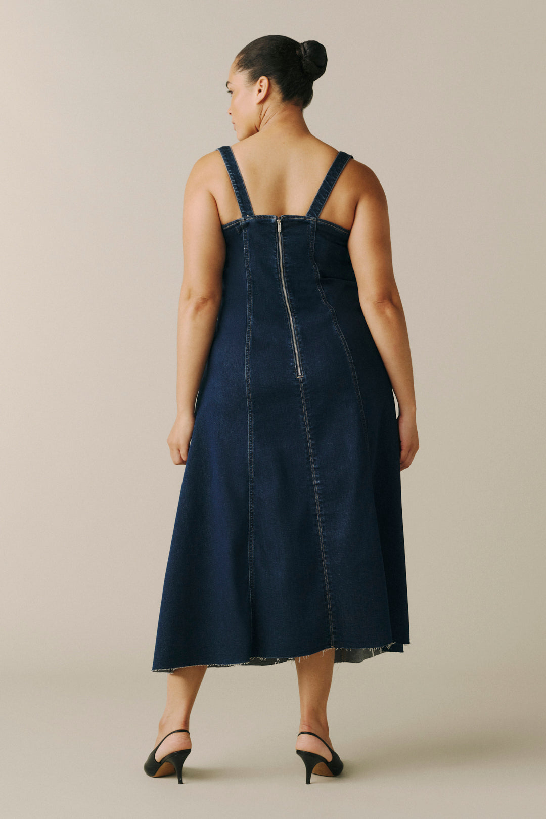 A woman shown from the back wearing a dark blue denim midi dress with wide shoulder straps and a central zipper.