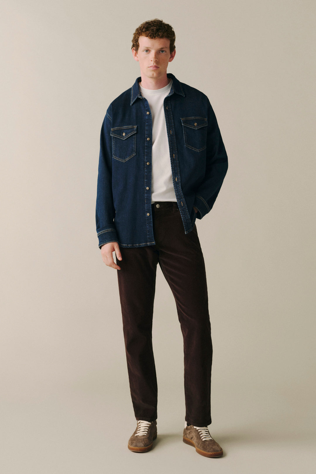 A man modeling a dark blue denim shirt jacket over a white t-shirt, paired with dark brown trousers and brown suede shoes, against a solid light beige background.