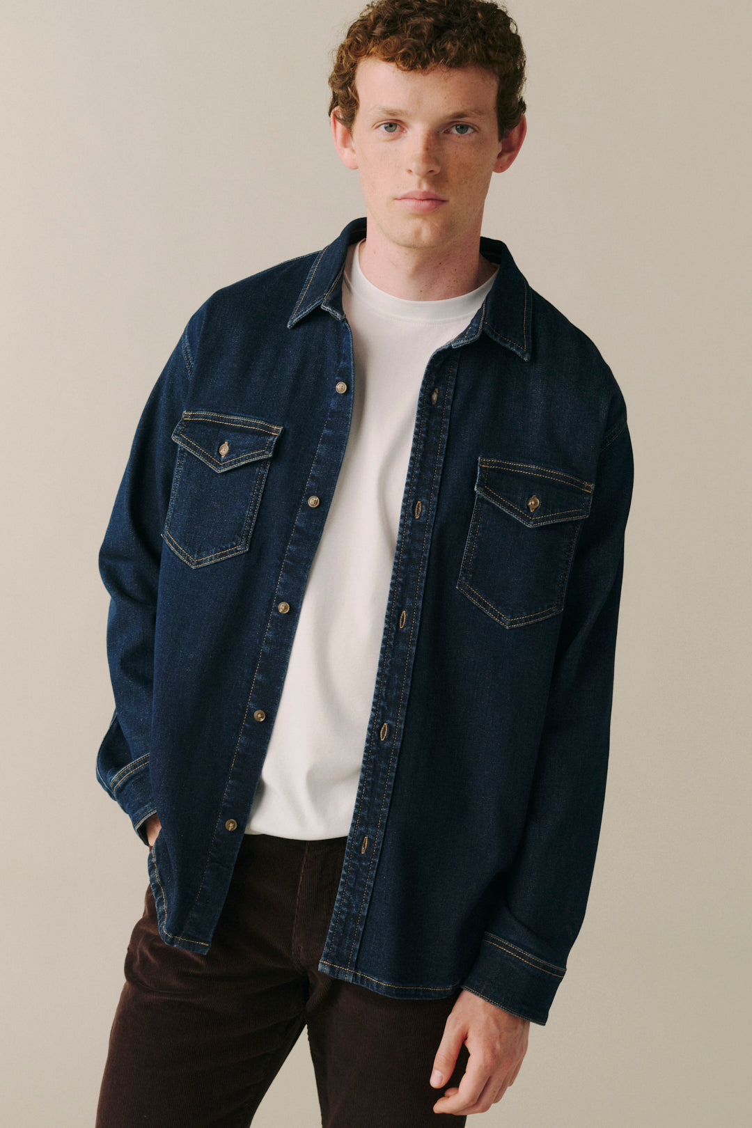 A man wearing a dark blue denim button-up shirt jacket over a white t-shirt and dark brown pants, standing against a plain beige studio background.