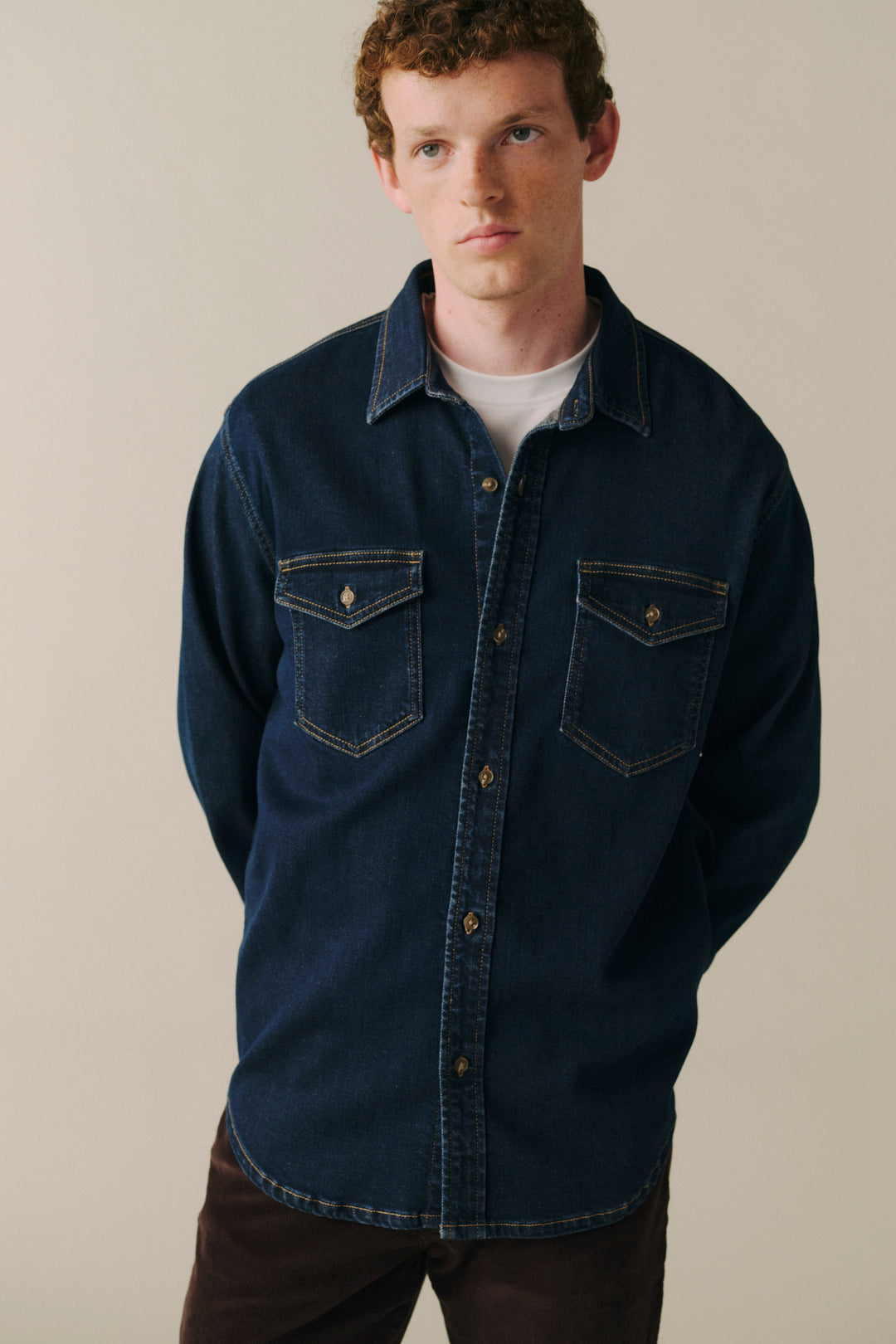 A man wearing a dark indigo denim button-up shirt with two chest pockets, layered over a white t-shirt and paired with brown pants, photographed against a plain beige studio background.