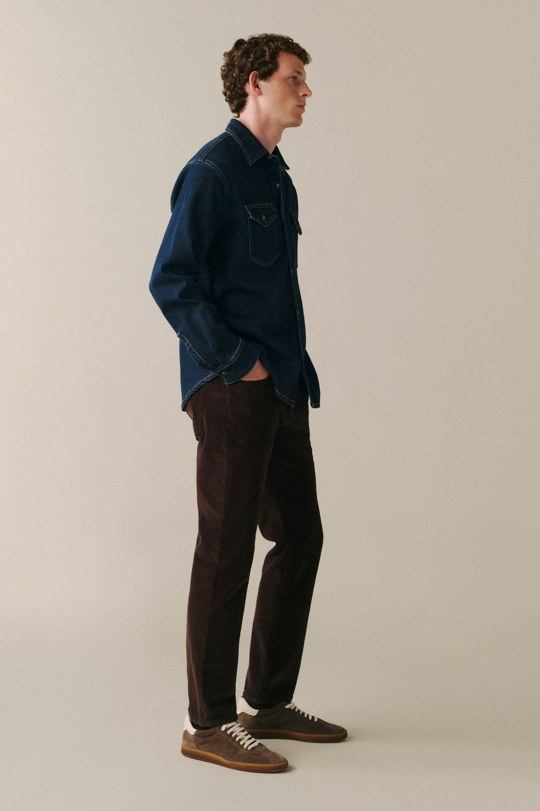 A man stands in profile wearing a dark navy denim shirt jacket, dark brown corduroy trousers, and brown suede sneakers against a plain light beige studio background.