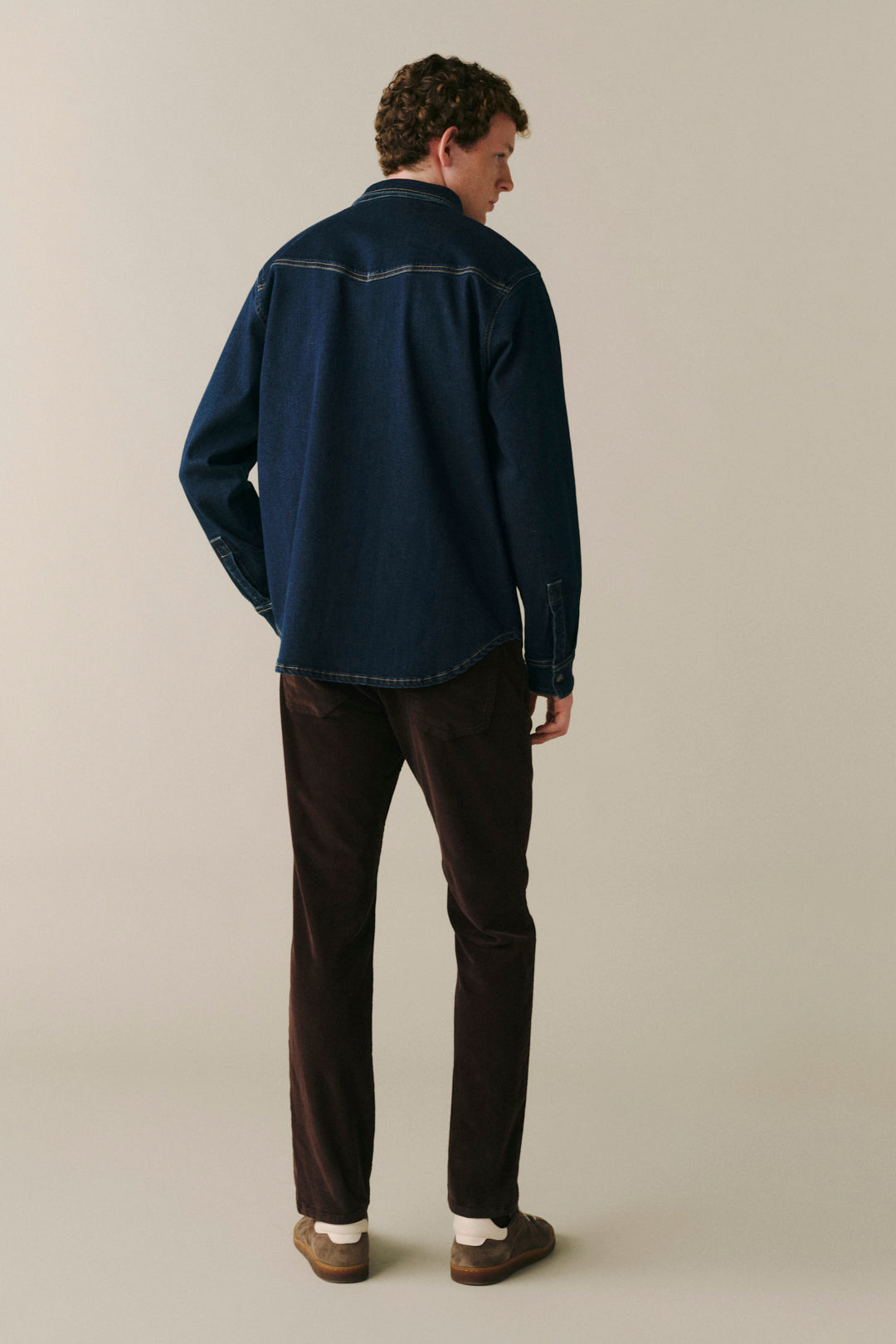 A man standing with his back to the camera wearing a dark blue denim shirt, dark brown corduroy pants, and brown shoes against a plain beige studio background.