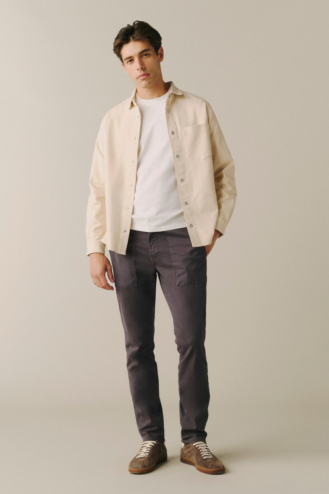 A man modeling a light beige overshirt over a white t-shirt, paired with dark charcoal gray pants and brown suede shoes, standing against a solid light beige background.