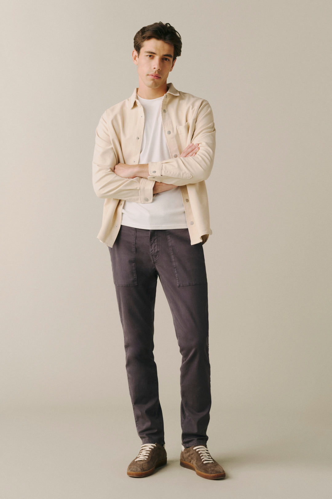A man stands with arms crossed wearing a light beige overshirt over a white t-shirt, dark charcoal pants, and brown suede shoes, photographed against a plain light beige studio background.
