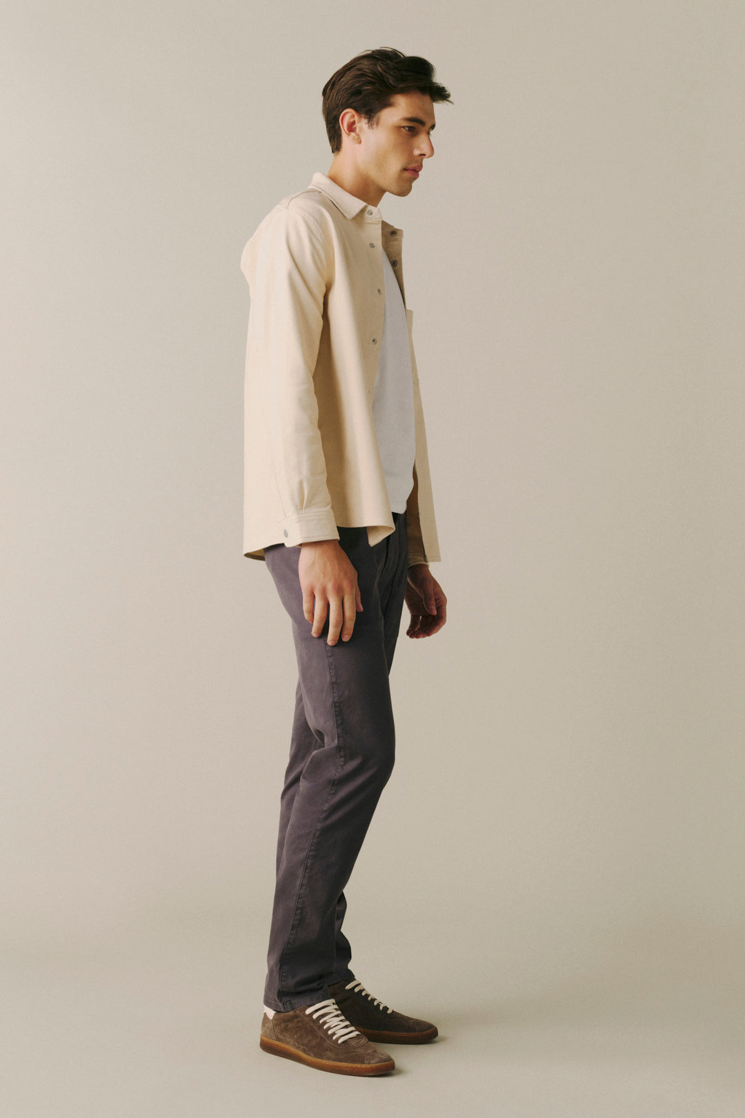 A man in a side profile wearing a cream button-down shirt over a white undershirt, dark gray trousers, and brown suede shoes, standing against a solid light beige studio background.