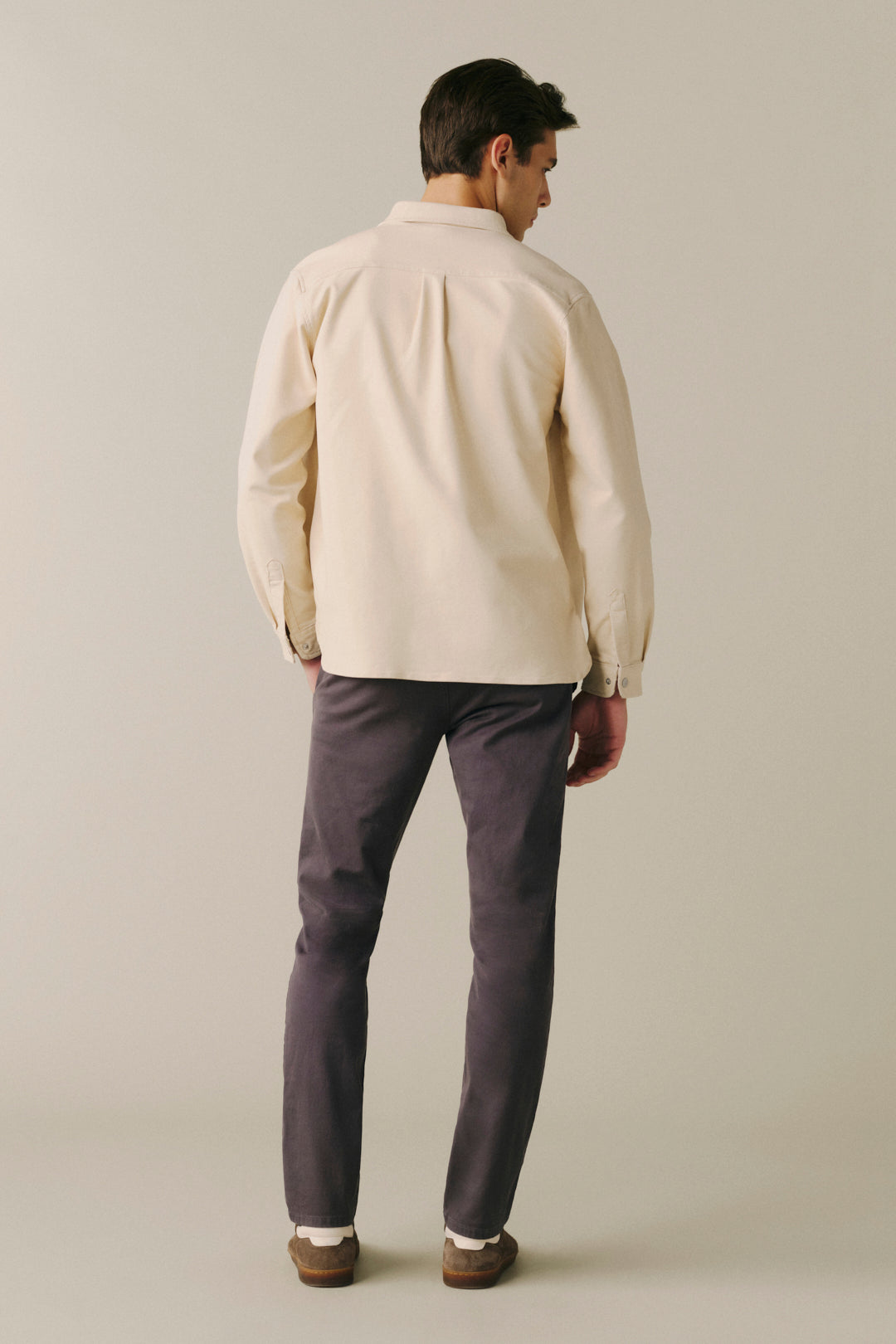 A man viewed from behind wearing a cream-colored long-sleeve button-down shirt and dark gray trousers, standing against a solid light beige studio background.