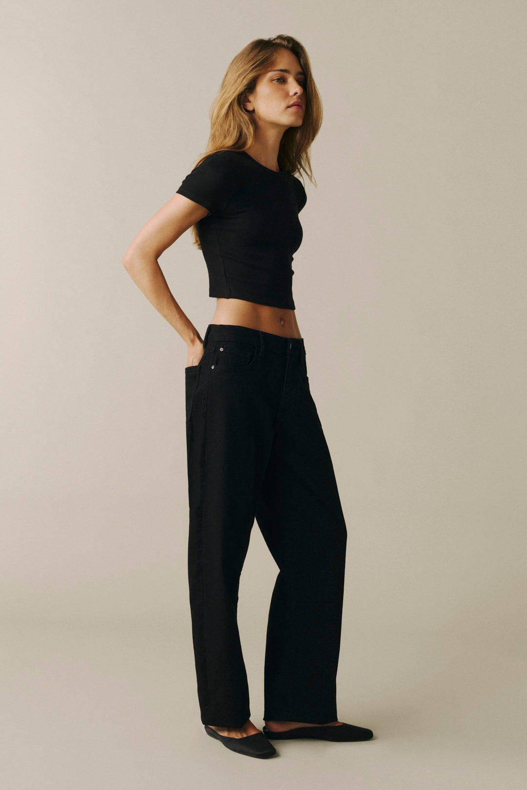 A woman modeling a black short-sleeve cropped t-shirt and black wide-leg trousers, standing in profile against a solid light beige studio background.
