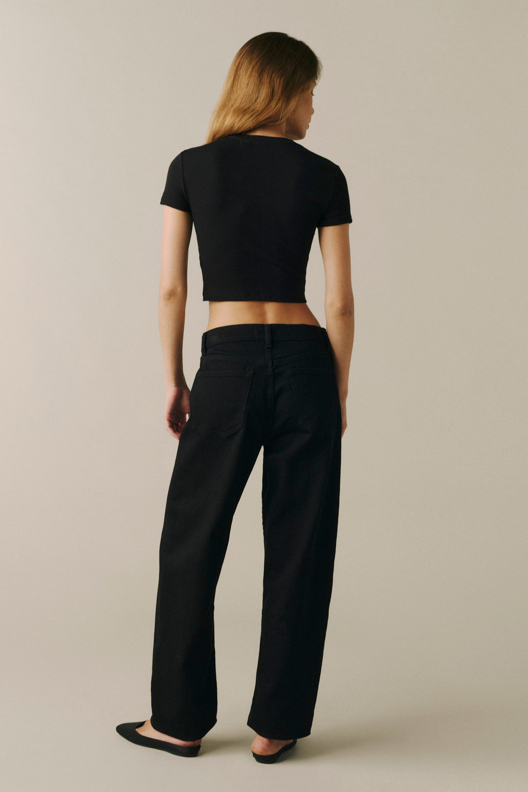 A woman is shown from behind wearing a black short-sleeve crop top and black wide-leg trousers, standing against a plain beige studio background.