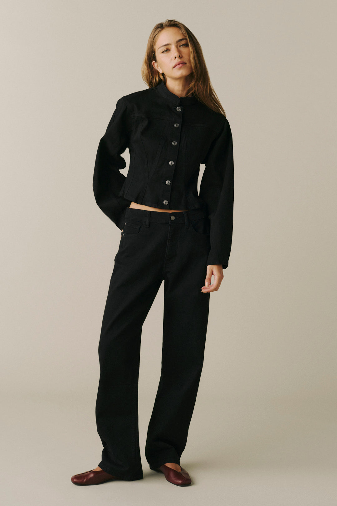 A woman modeling a black cropped jacket with a high mock neck and front buttons, paired with black wide-leg trousers and dark brown loafers, against a solid light beige studio background.
