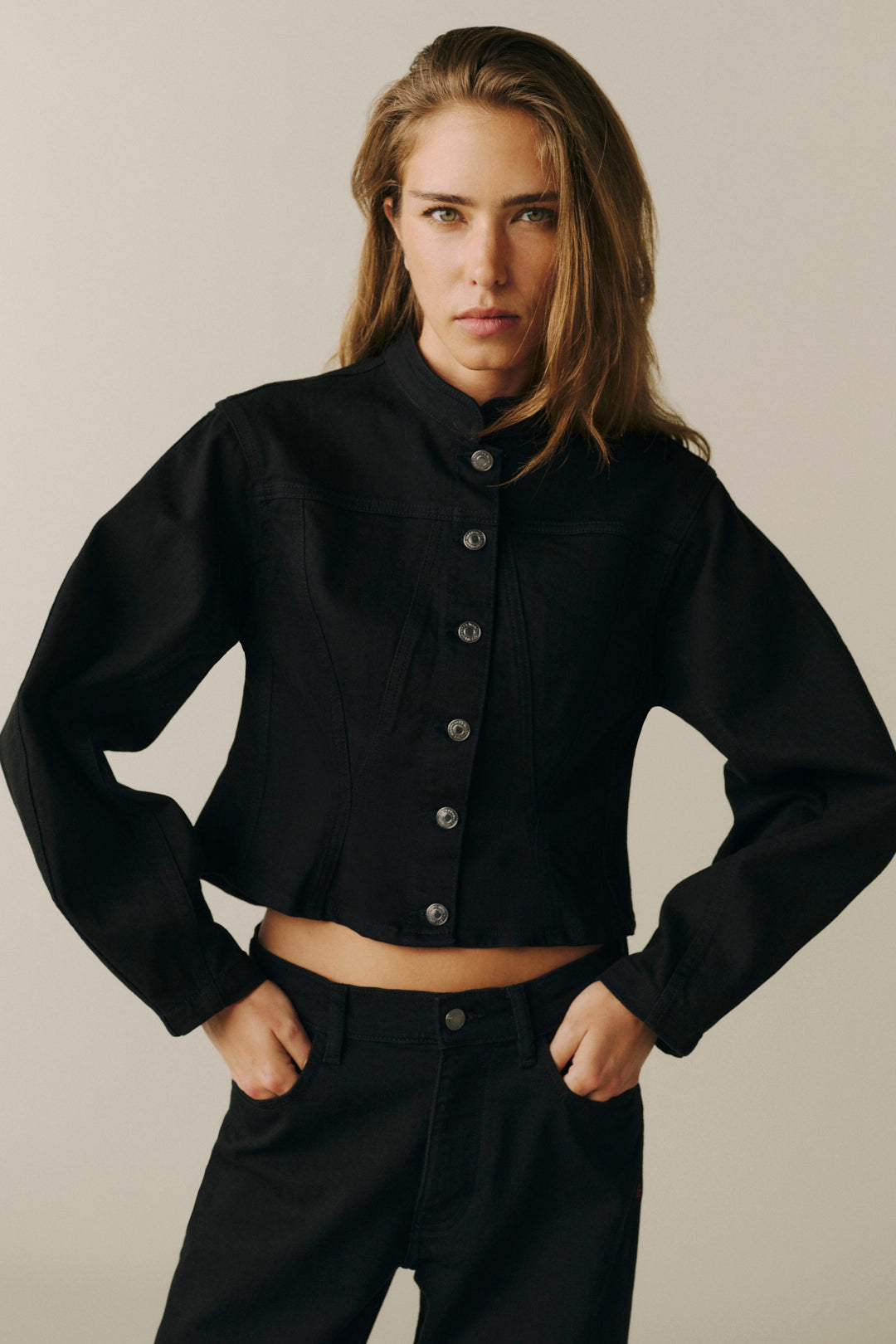 A woman modeling a black cropped button-front jacket with a stand collar and black pants against a plain light beige studio background.