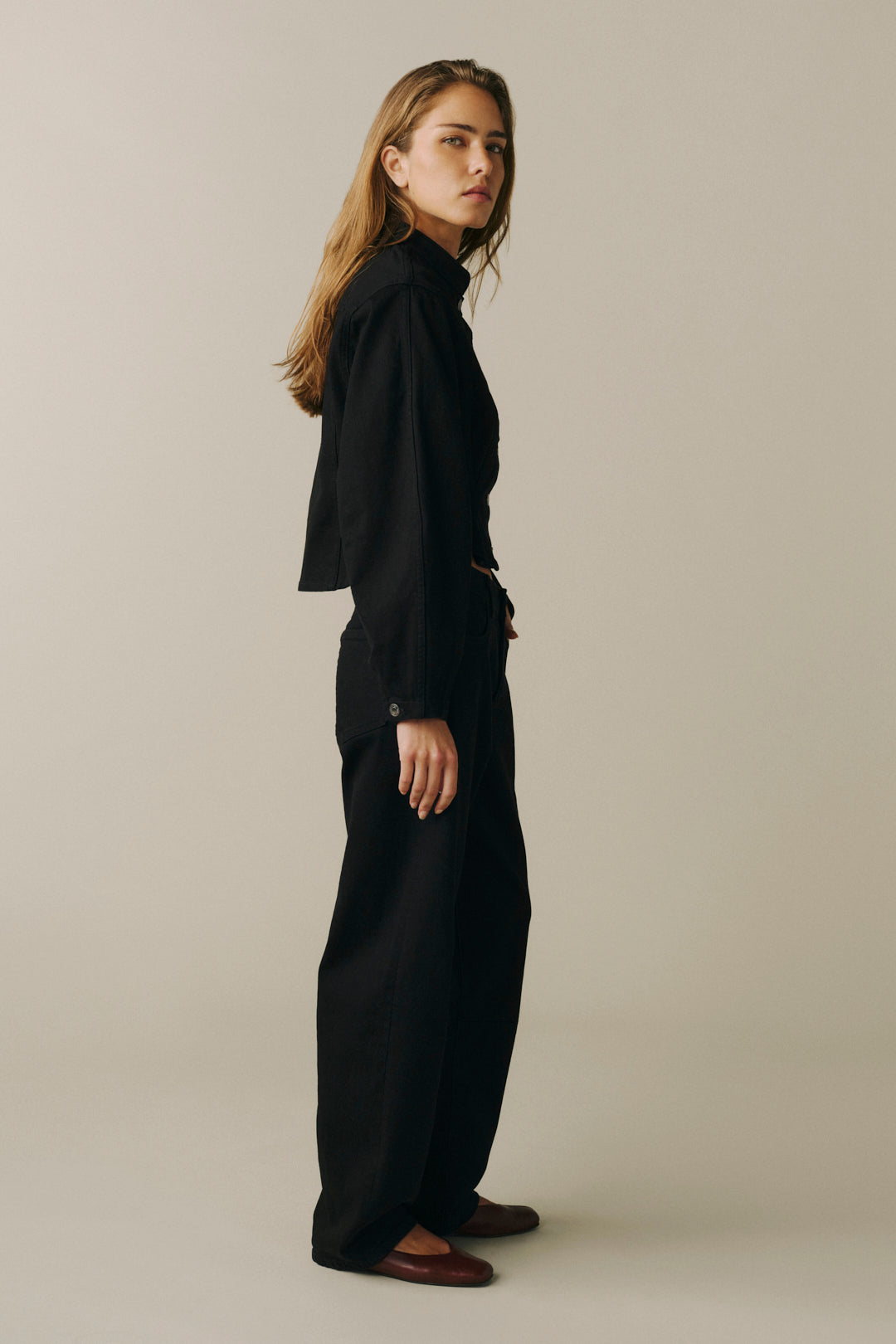 A woman stands in profile wearing a black long-sleeve high-neck top and matching black wide-leg trousers.